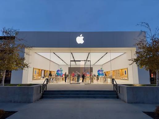 Filas por el iPhone Duo en Londres, Macao y Sídney Fachada de vidrio de una tienda Apple iluminada al atardecer, con clientes en su interior